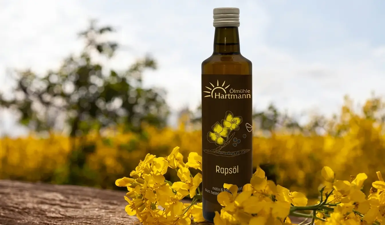 Basisöle | Ölmühle Hartmann Eine Flasche Rapsöl steht auf einem Tisch, neben einer Rapspflanze und im Hintergrund ist ein Rapsfeld